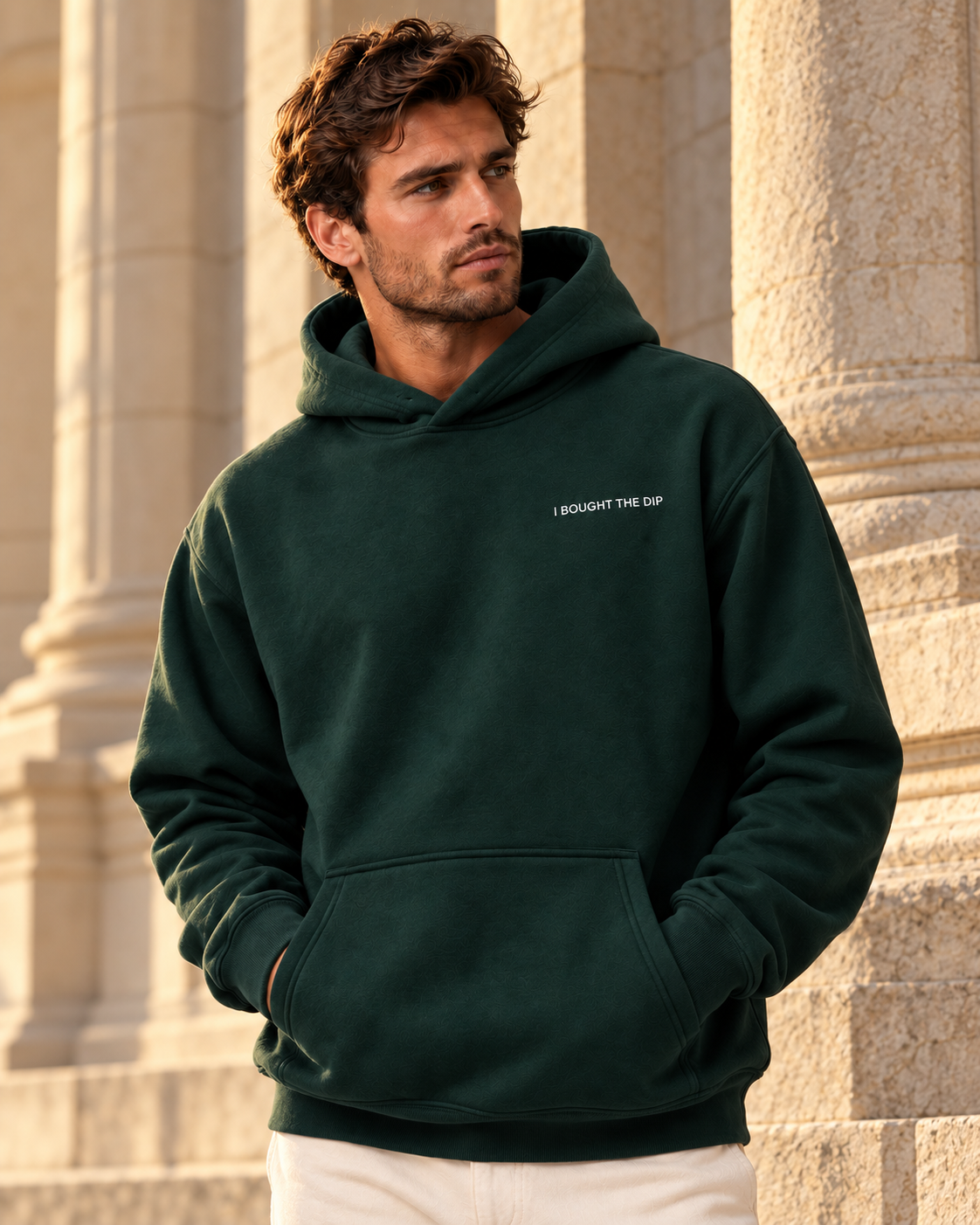 Man wearing the Pine Green I Bought The Dip hoodie with white left-chest embroidery beside pale stone columns in warm afternoon light.